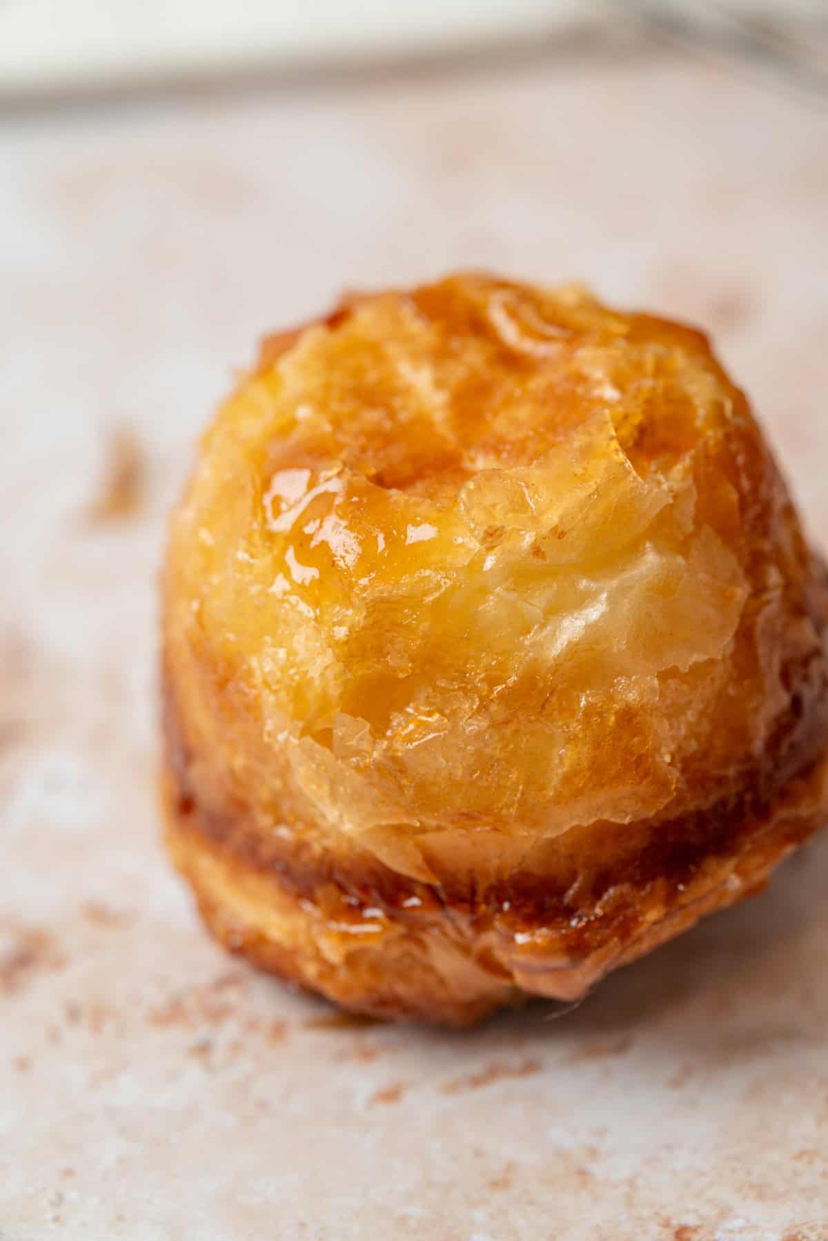 A close-up of a golden-brown, glossy sourdough kouign amann pastry with flaky layers, sitting on a light-colored surface. The pastry appears caramelized and slightly shiny, suggesting a sweet glaze or syrup.