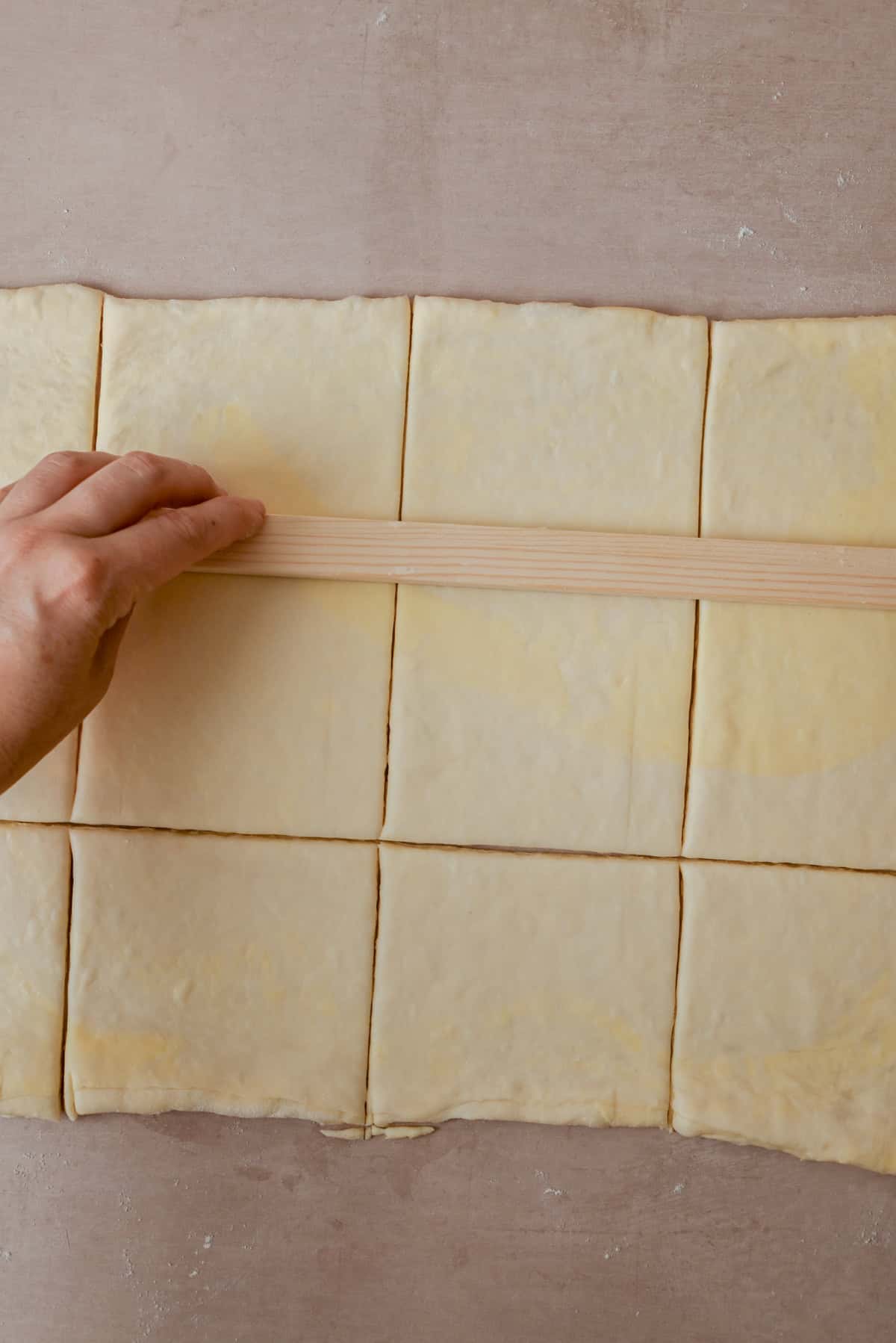 A hand uses a wooden ruler to measure and cut a sheet of dough into evenly sized rectangular pieces on a lightly floured surface.
