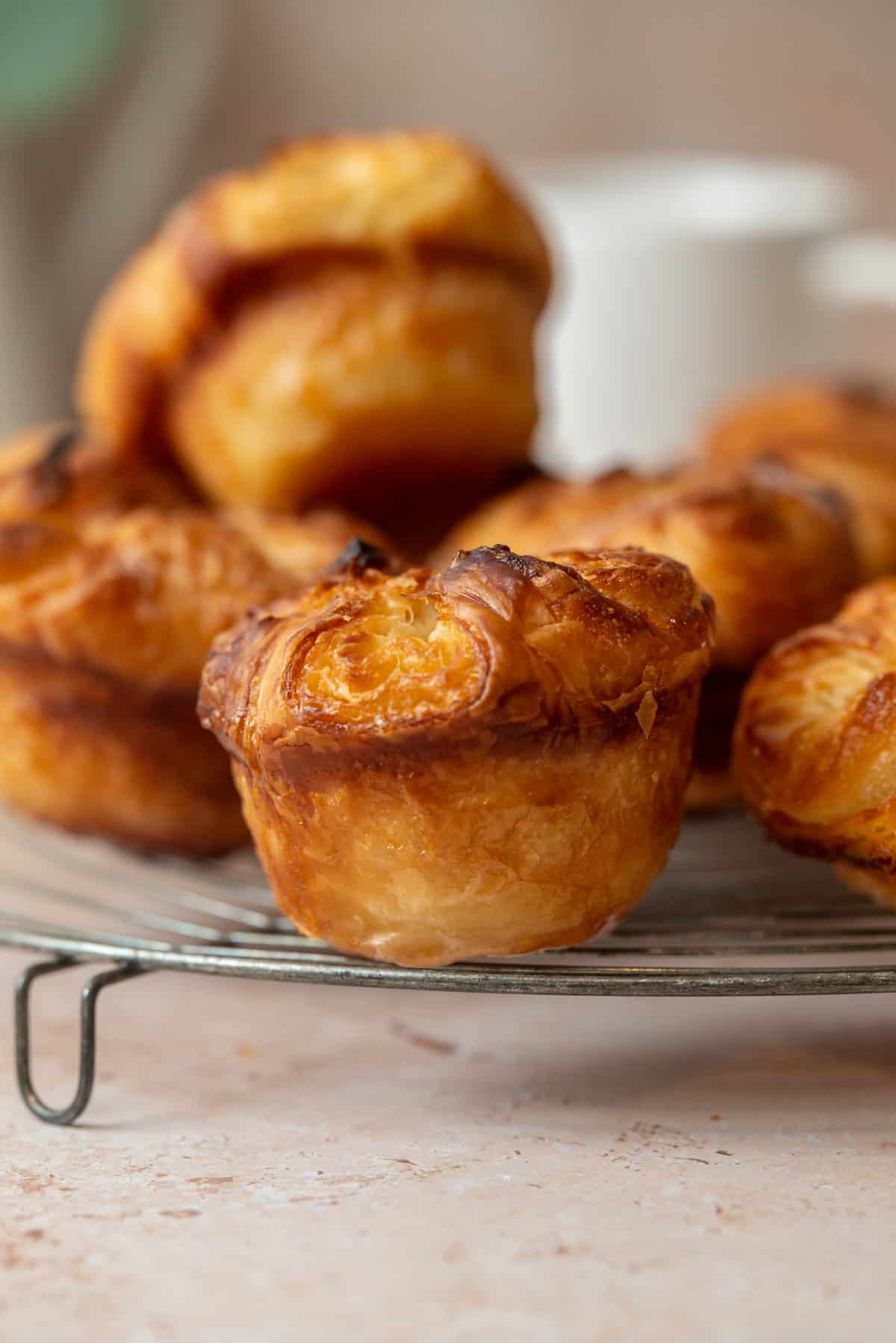 A close-up of golden, flaky sourdough kouign amann pastries arranged on a round wire cooling rack, with a blurred cup and more pastries in the background.