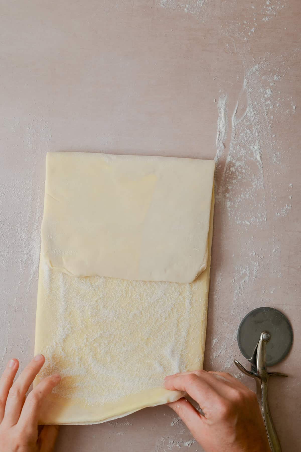 Hands folding a sheet of dough sprinkled with flour on a light surface, with a fluted pastry wheel cutter lying nearby.