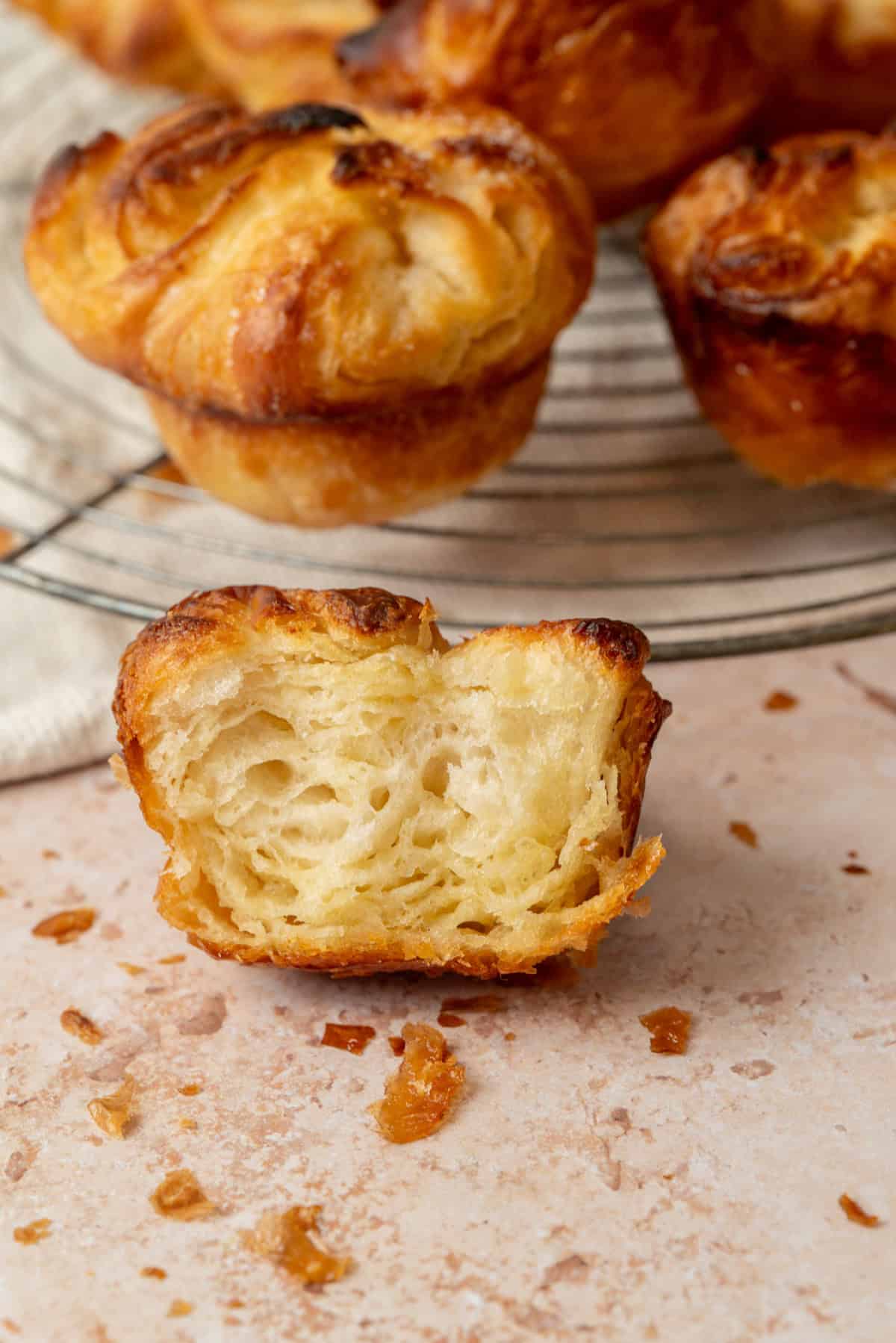 A close-up of a golden, flaky pastry with a bite taken out, showing its airy, layered interior. More similar pastries are visible on a wire rack in the background, with scattered crumbs on the surface.