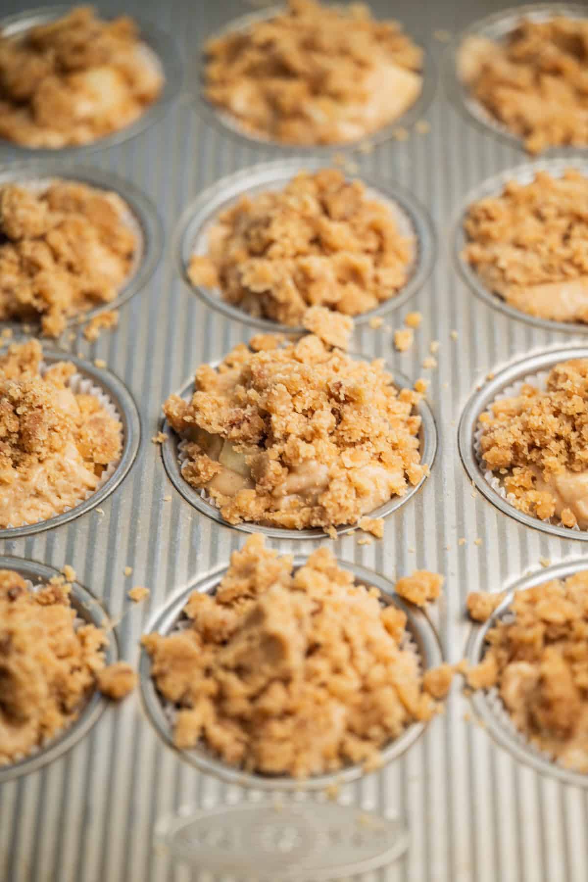A close-up view of a metal muffin tin filled with unbaked muffins topped with a crumbly streusel topping, ready to be baked.
