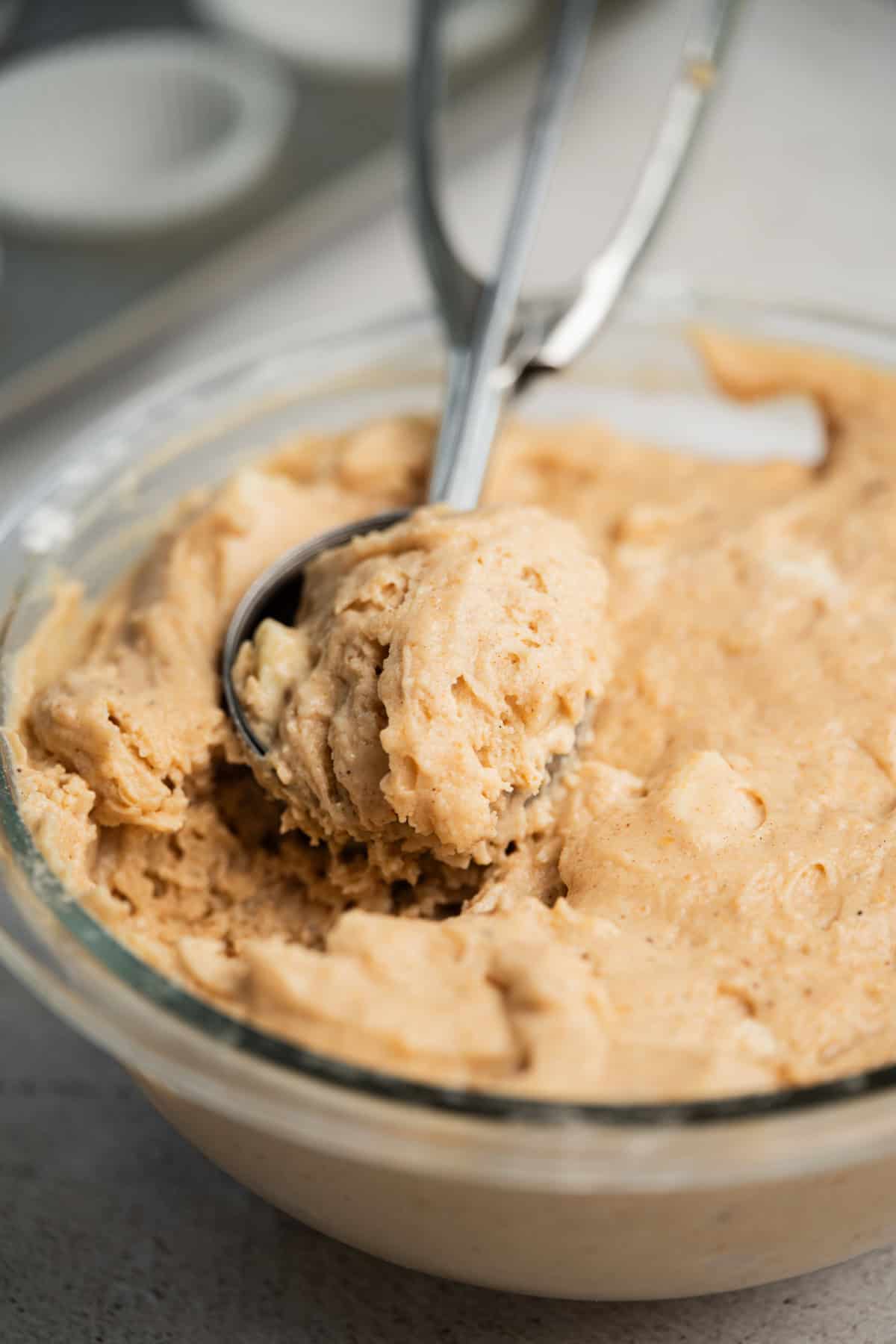 A metal ice cream scoop is lifting thick, light brown muffin or cookie dough from a glass mixing bowl. A muffin tin with paper liners is blurred in the background.