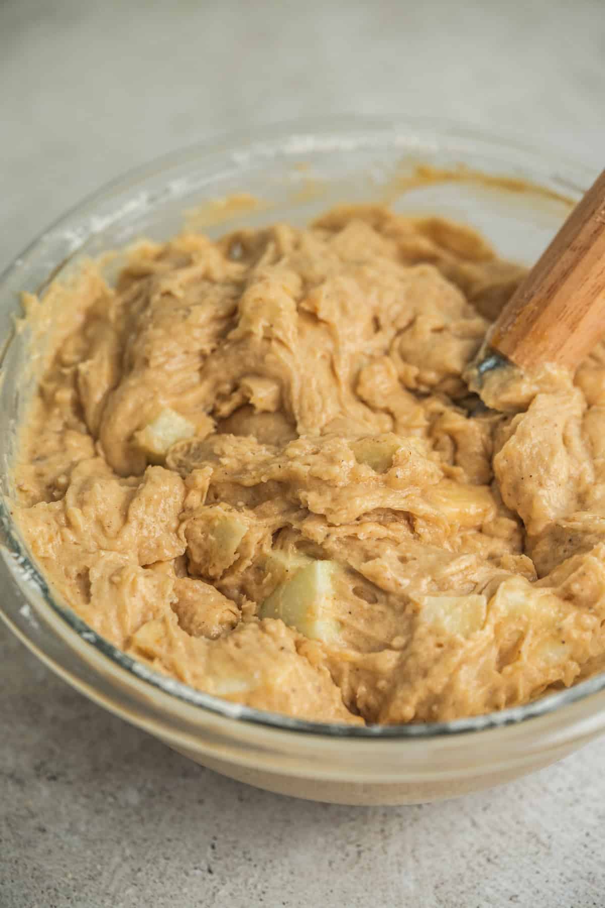 A glass bowl filled with thick, light brown batter and visible chunks, being mixed with a wooden spatula on a light surface.