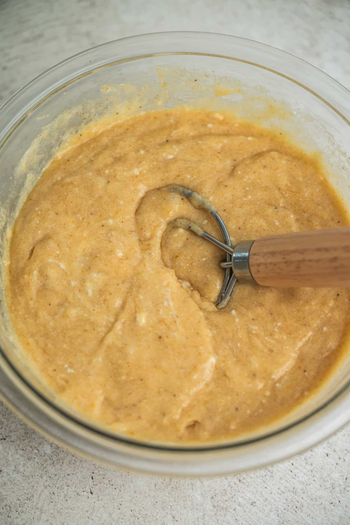 A glass bowl filled with thick, light brown batter being mixed with a wooden-handled whisk, resting on a light-colored countertop.
