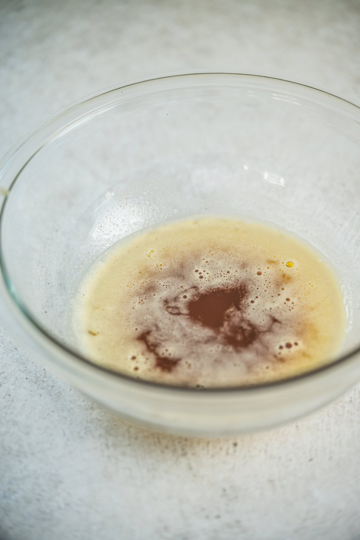 A clear glass bowl containing melted butter with a swirl of vanilla extract on top, sitting on a light, textured surface.