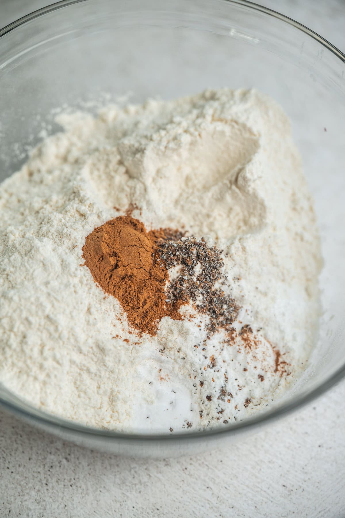 A glass bowl filled with flour, ground cinnamon, ground cardamom, and baking powder, ready to be mixed for baking.