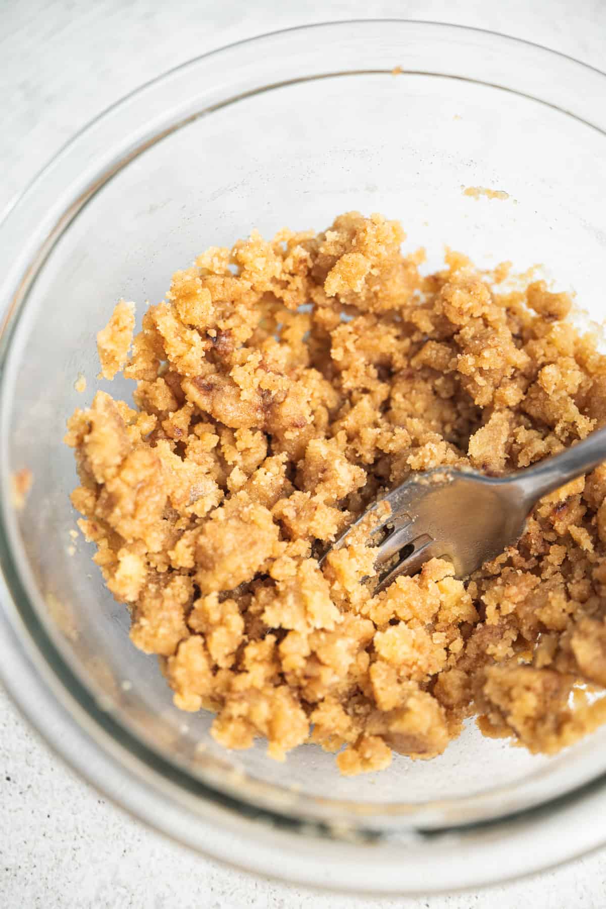 A glass bowl contains a crumbly, light brown mixture with a metal fork partially submerged, indicating the mixture is being stirred or mashed. The bowl is on a light-colored surface.