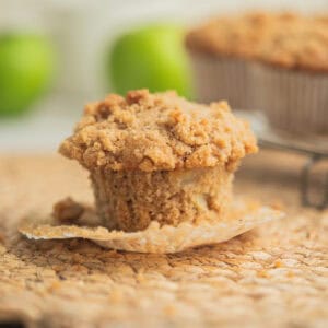 A close-up of a crumb-topped apple muffin with its paper wrapper partially peeled away, sitting on a woven placemat. Green apples and more muffins appear blurred in the background.