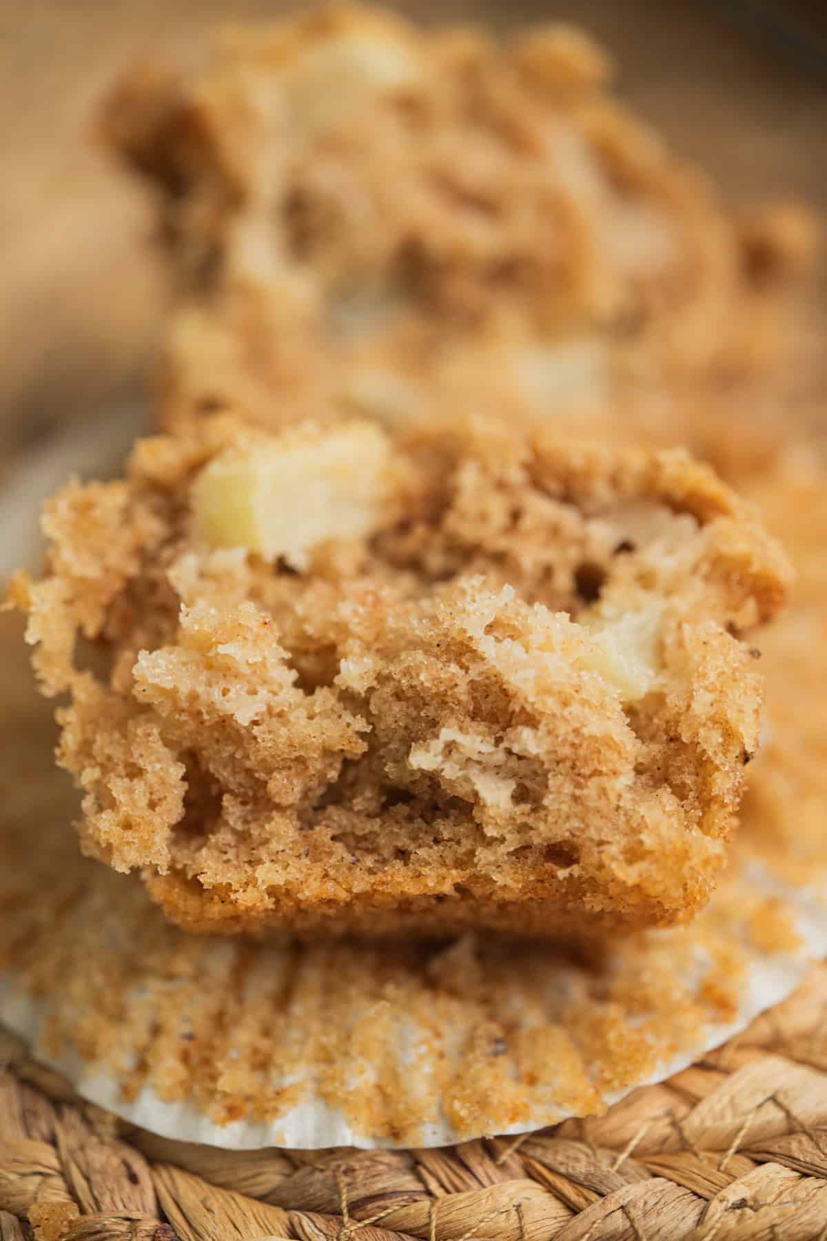 A close-up of a halved muffin with a moist, crumbly texture and visible chunks of apple, resting on a white paper muffin liner. The background is softly blurred.