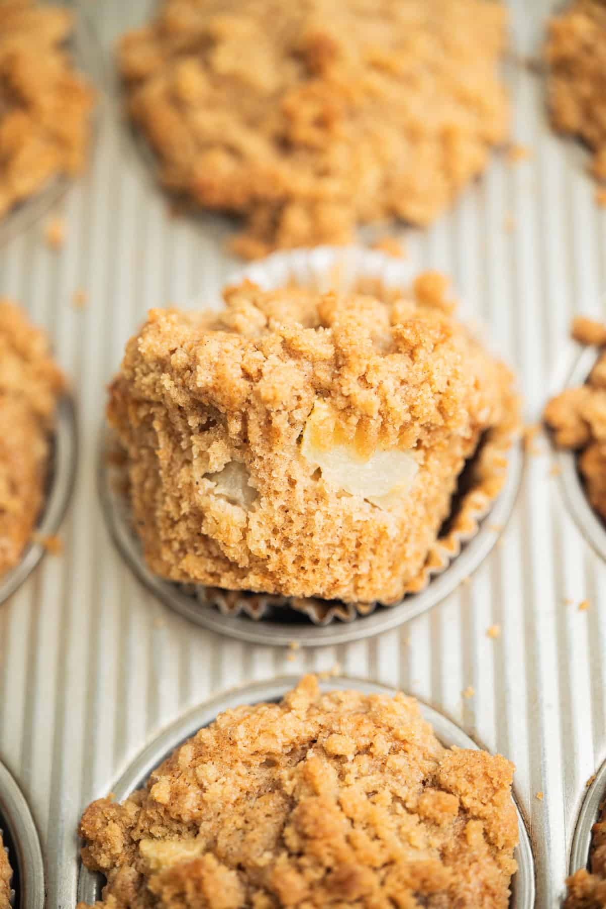 A close-up of a muffin with a crumbly streusel topping in a muffin tin. The muffin is partially unwrapped, revealing chunks of fruit inside and a golden-brown, textured surface.