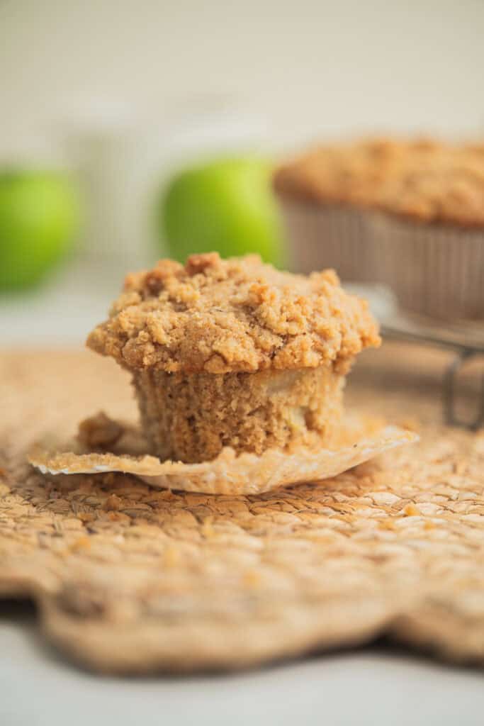 A close-up of a crumb-topped apple muffin with its paper wrapper partially peeled away, sitting on a woven placemat. Green apples and more muffins appear blurred in the background.
