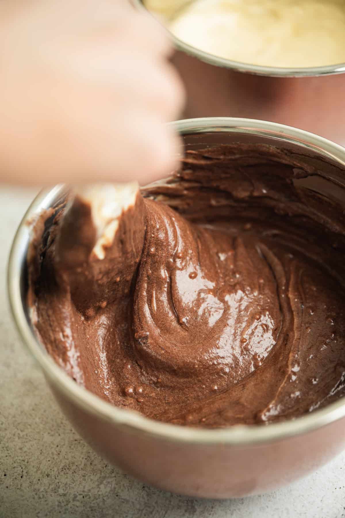 A hand uses a spatula to mix smooth, glossy chocolate batter in a metal bowl, with another bowl containing light batter in the background.