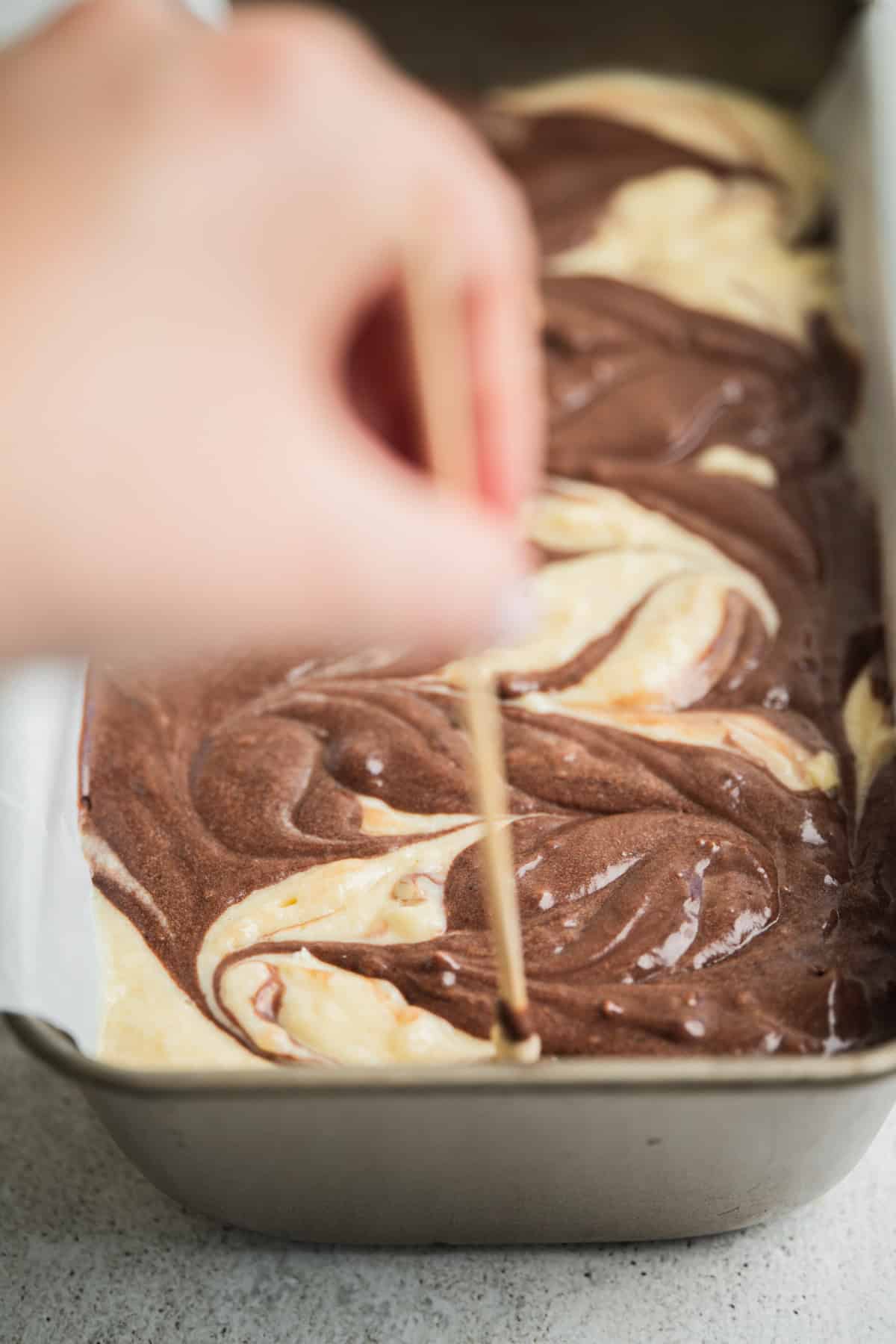A hand swirls a skewer through chocolate and vanilla cake batter in a parchment-lined loaf pan, creating a marbled pattern before baking.
