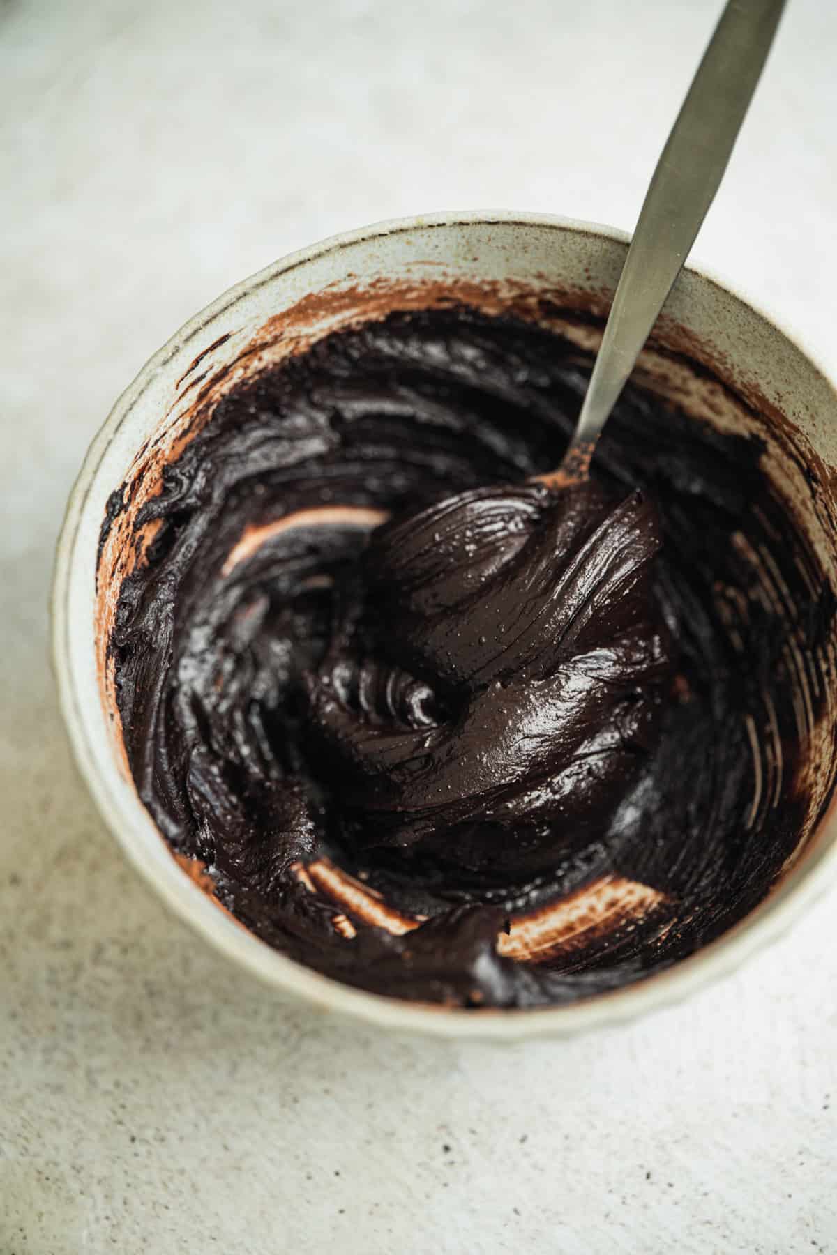 A bowl of thick, glossy chocolate batter being mixed with a spoon on a light-colored surface.