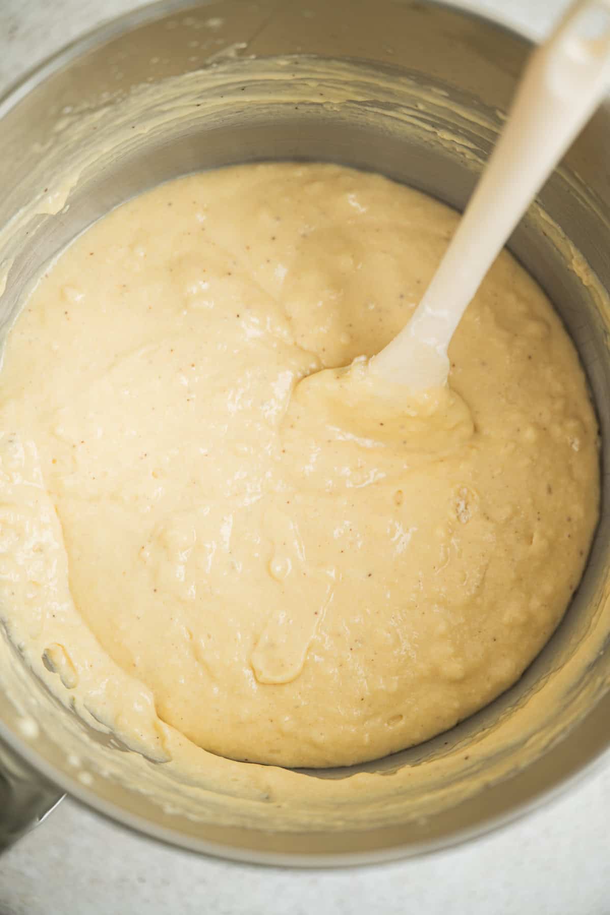 A large metal mixing bowl filled with creamy, pale yellow batter. A white spatula is partially submerged in the batter, showing it has been mixed to a smooth consistency.