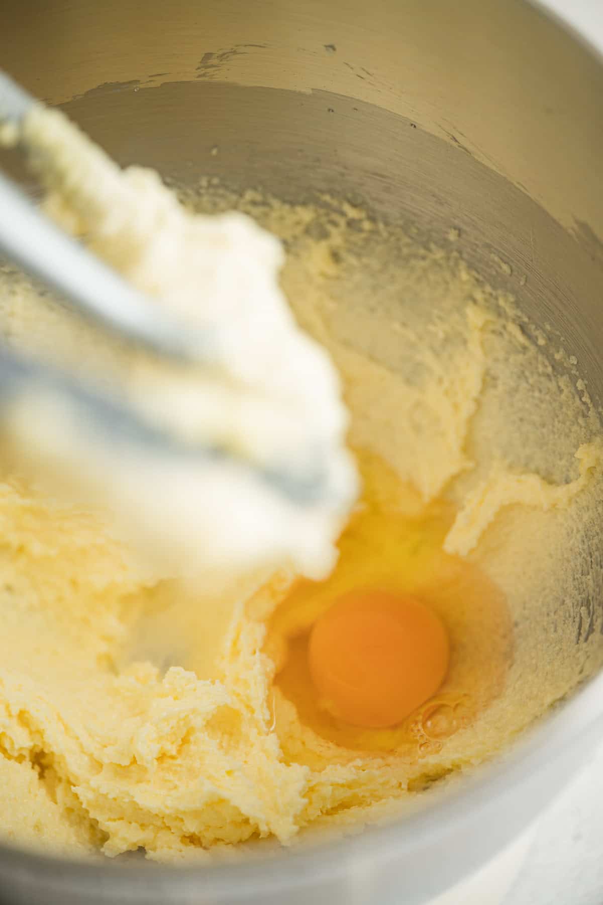 A close-up of an egg cracked into a mixing bowl with creamed butter and sugar, with a metal beater partially visible in the upper left corner.