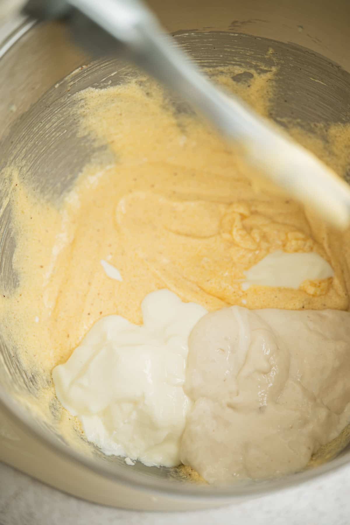 A metal mixing bowl contains a yellow batter, with dollops of white yogurt and a beige mixture on top, ready to be combined with a spatula.
