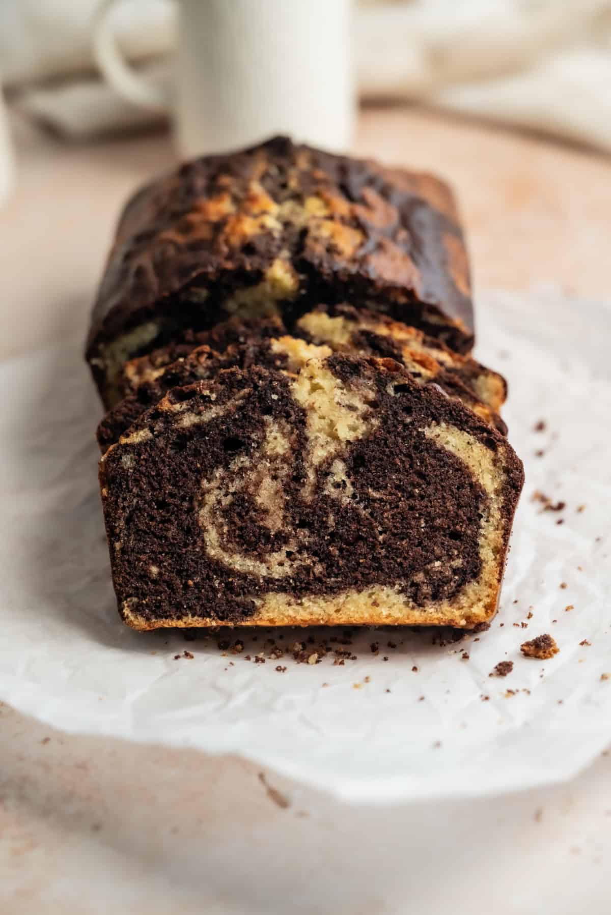A sliced loaf of sourdough discard marble cake with swirls of chocolate and vanilla on white parchment paper, with a blurred white mug in the background.