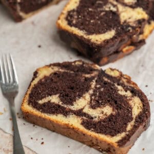 A slice of sourdough marble cake with swirls of chocolate and vanilla sits on parchment paper next to a silver fork, with more slices of cake in the background.