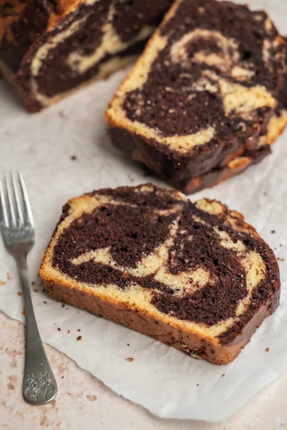 A slice of sourdough marble cake with swirls of chocolate and vanilla sits on parchment paper next to a silver fork, with more slices of cake in the background.