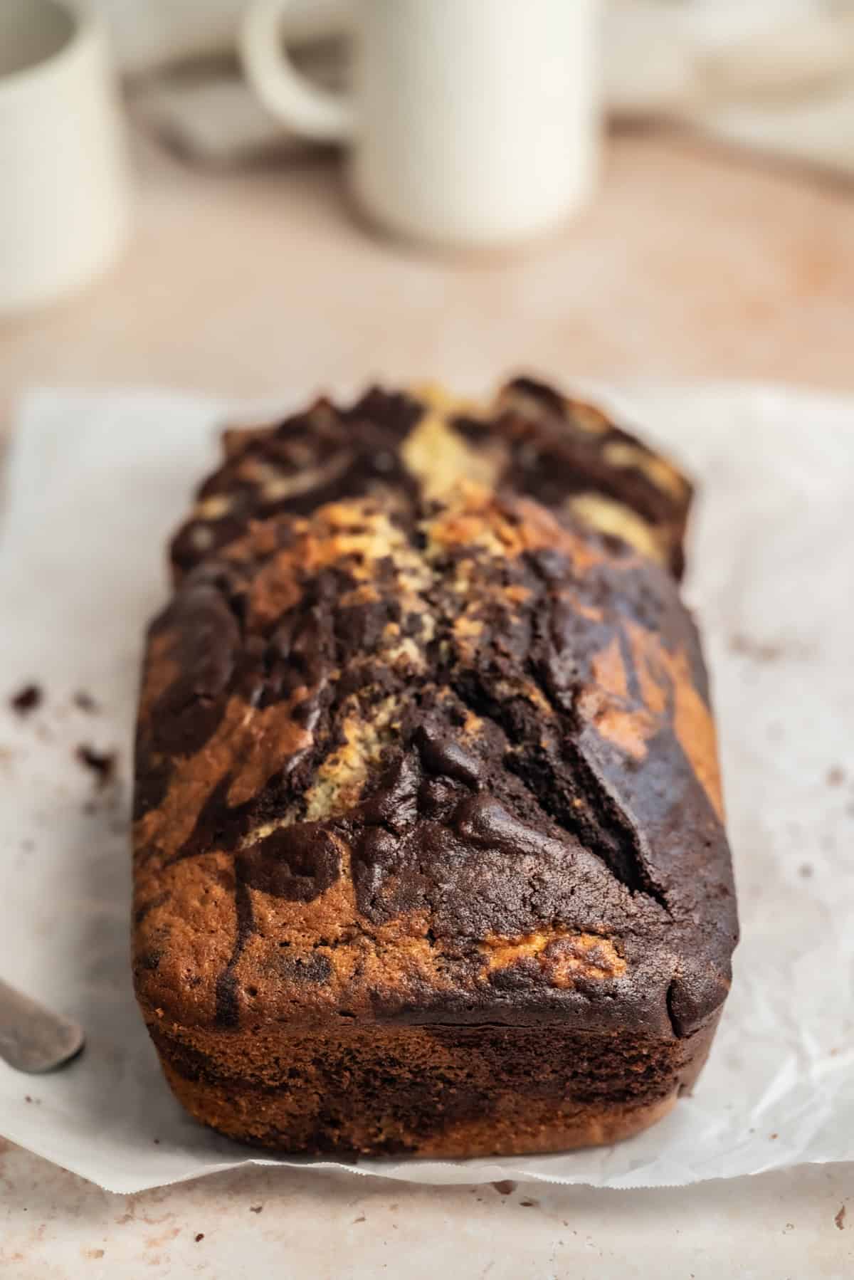 A freshly baked sourdough marble loaf cake with swirls of chocolate and vanilla, resting on a piece of parchment paper on a light countertop. Blurry white mugs are visible in the background.