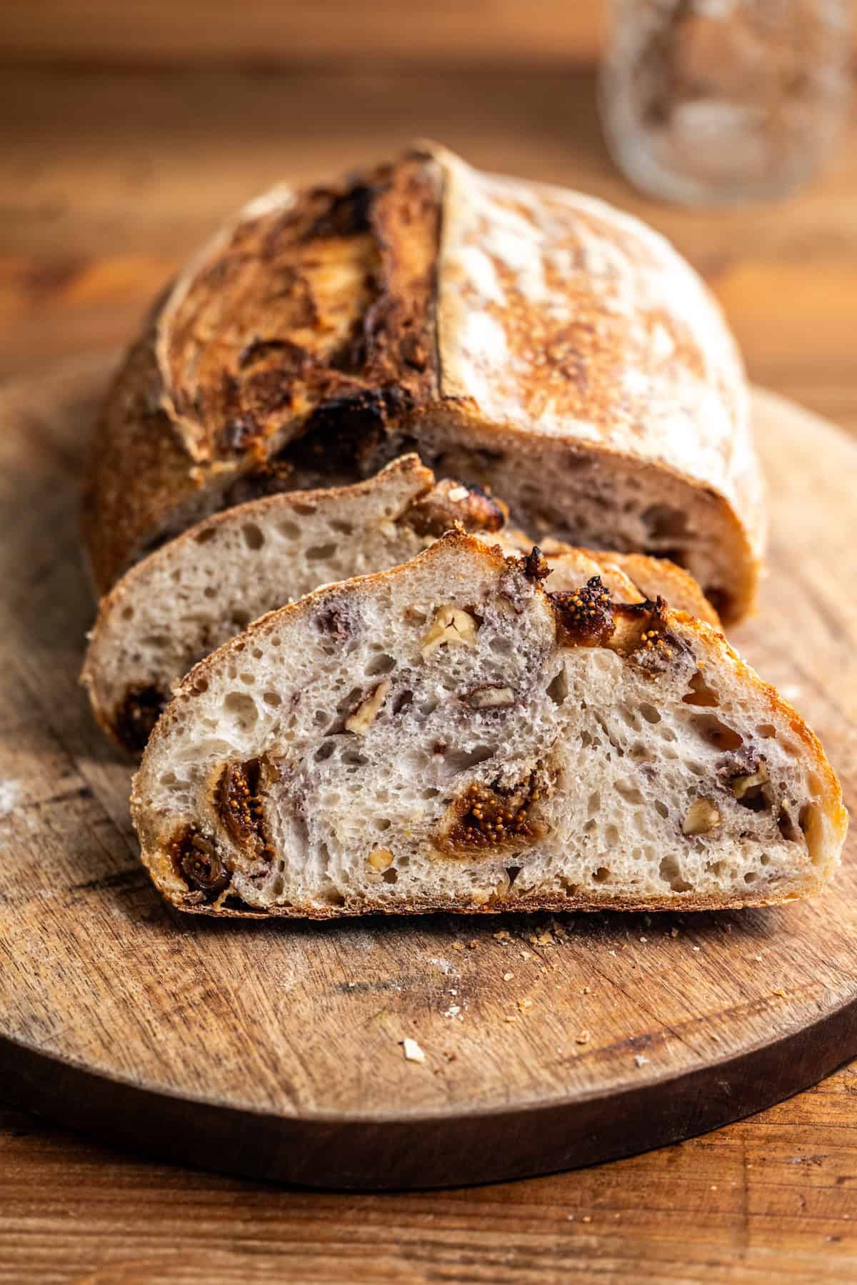 A sliced loaf of artisanal bread with a golden crust and a textured interior featuring pieces of nuts and dried fruit, displayed on a wooden cutting board.