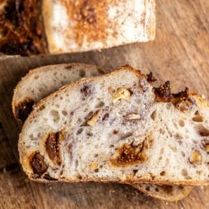 Sliced loaf of rustic bread with visible chunks of figs and walnuts on a wooden cutting board.