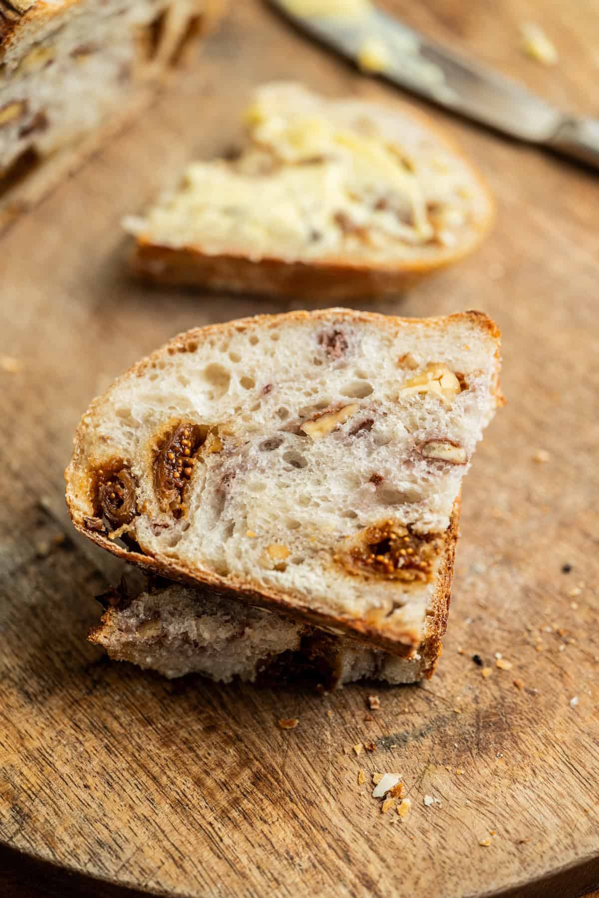Two slices of rustic bread with visible figs and nuts on a wooden cutting board. One slice in the background is spread with butter; a butter knife rests nearby.