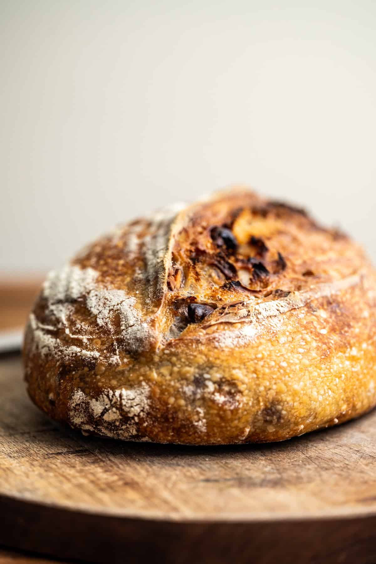 A round loaf of rustic artisan bread with a golden-brown crust sits on a wooden cutting board. The surface is textured with cracks and flour dusting.