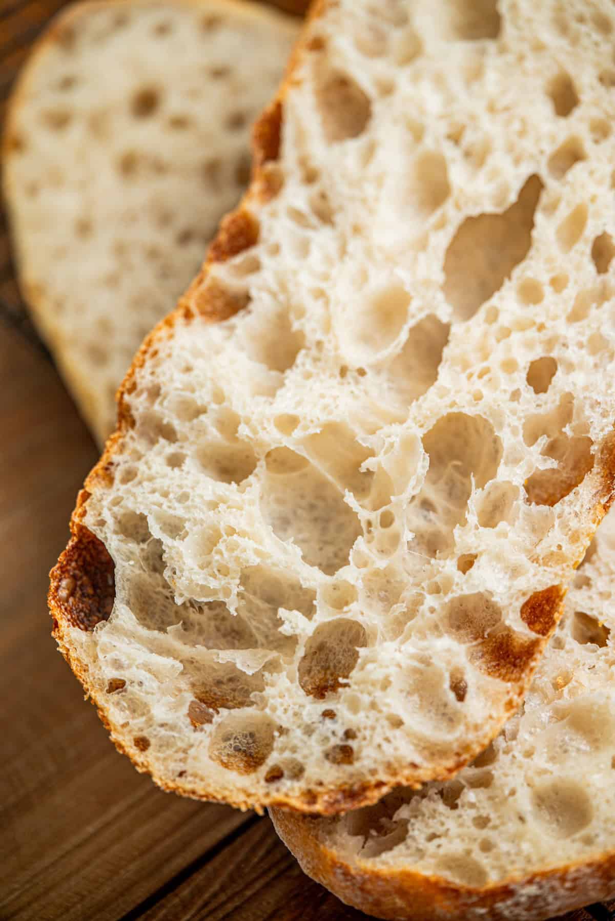 Close-up of two slices of rustic bread with a crispy crust and an airy, open crumb structure, placed on a wooden surface.