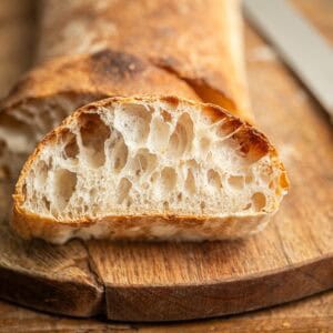 A sliced loaf of rustic bread with an airy, open crumb sits on a wooden cutting board beside a bread knife. The crust is golden brown and the bread appears freshly baked.