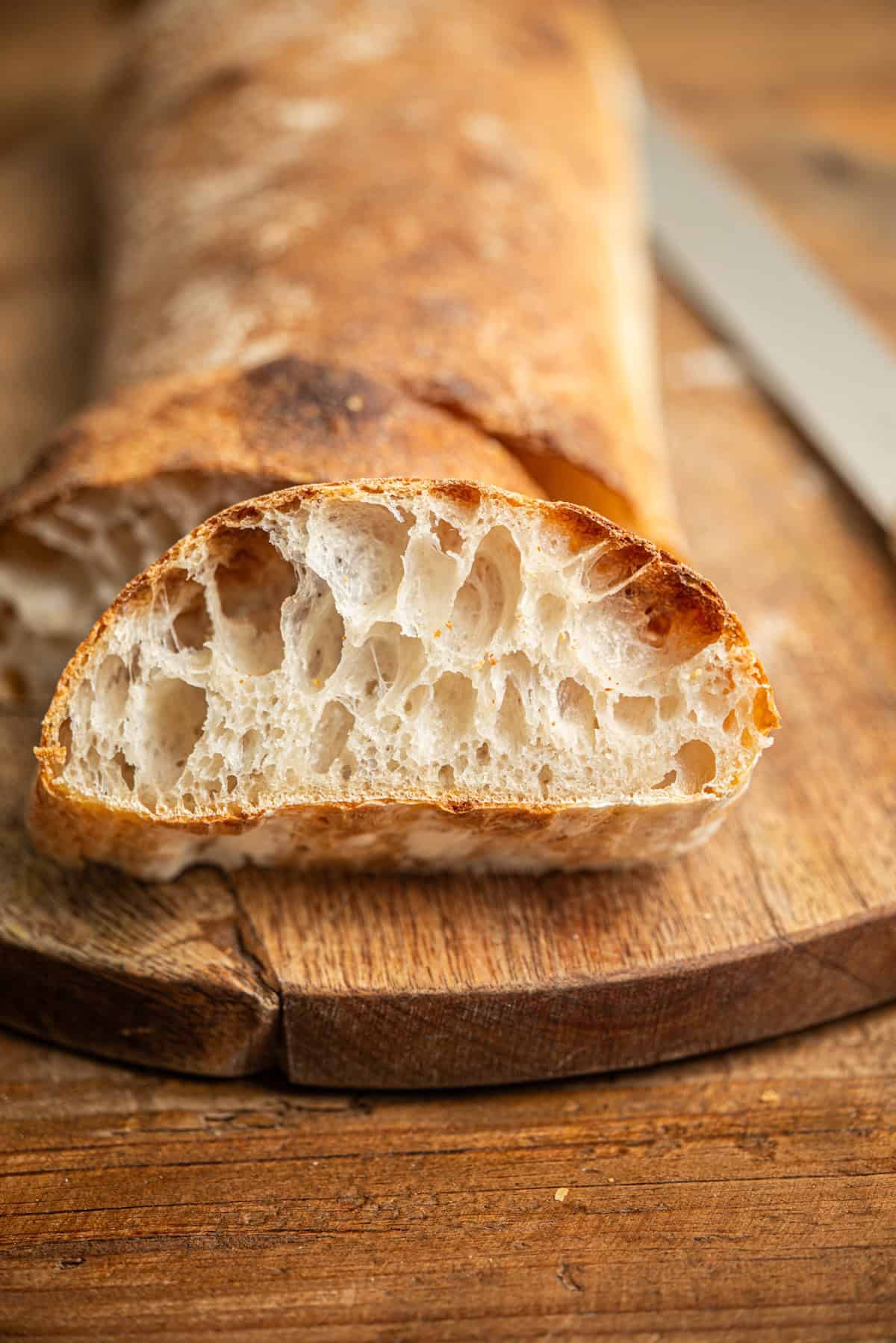 A sliced loaf of rustic bread with an airy, open crumb sits on a wooden cutting board beside a bread knife. The crust is golden brown and the bread appears freshly baked.