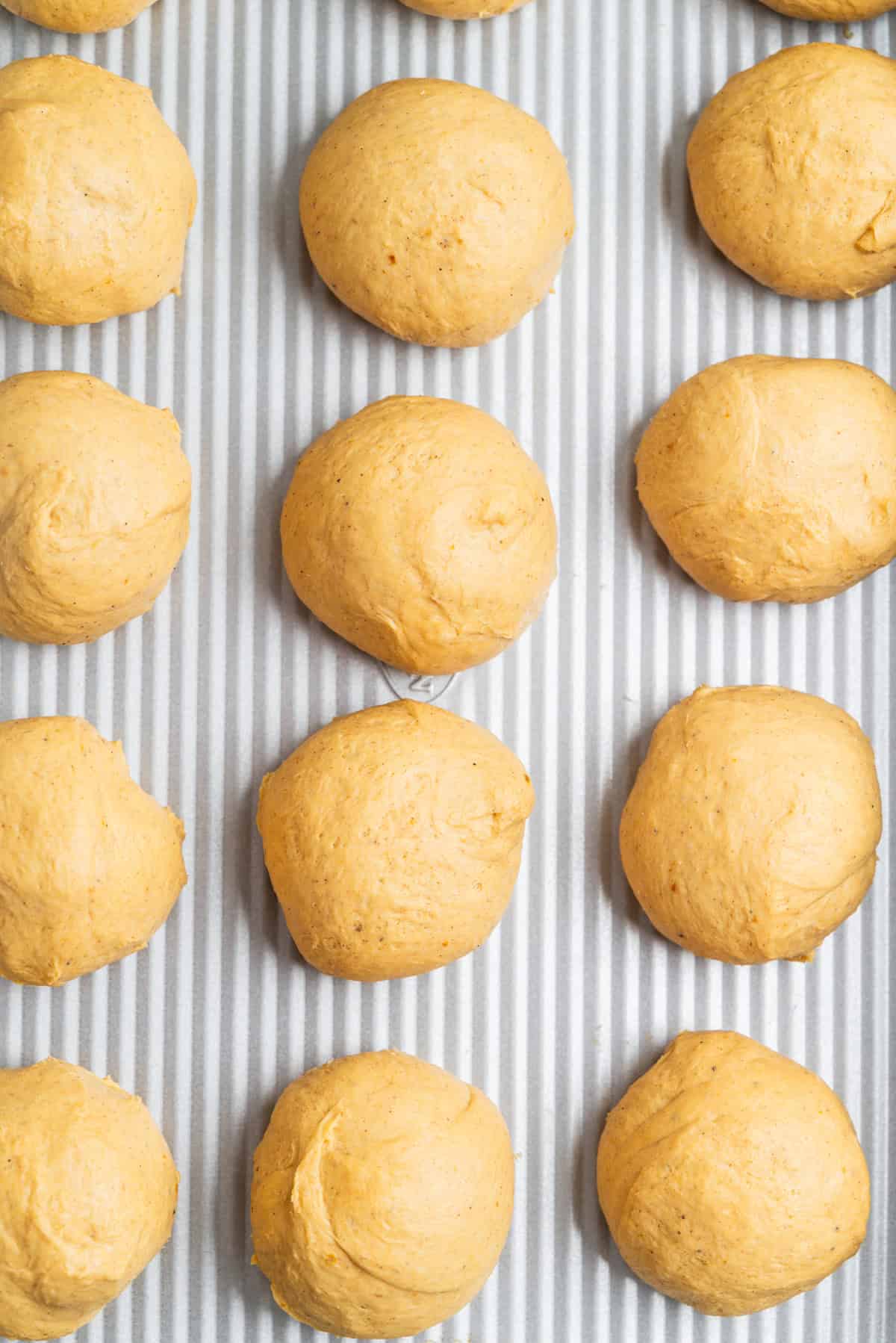 Round balls of dough arranged in neat rows on a lined baking sheet, ready to be baked. The dough is a light golden-brown color and the surface is slightly textured.