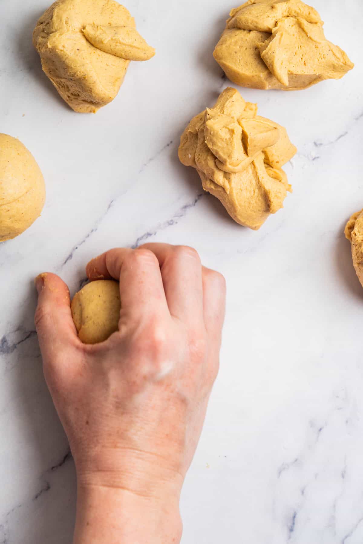 A hand shaping a ball of dough on a white marble surface, with several unshaped and shaped dough portions nearby.