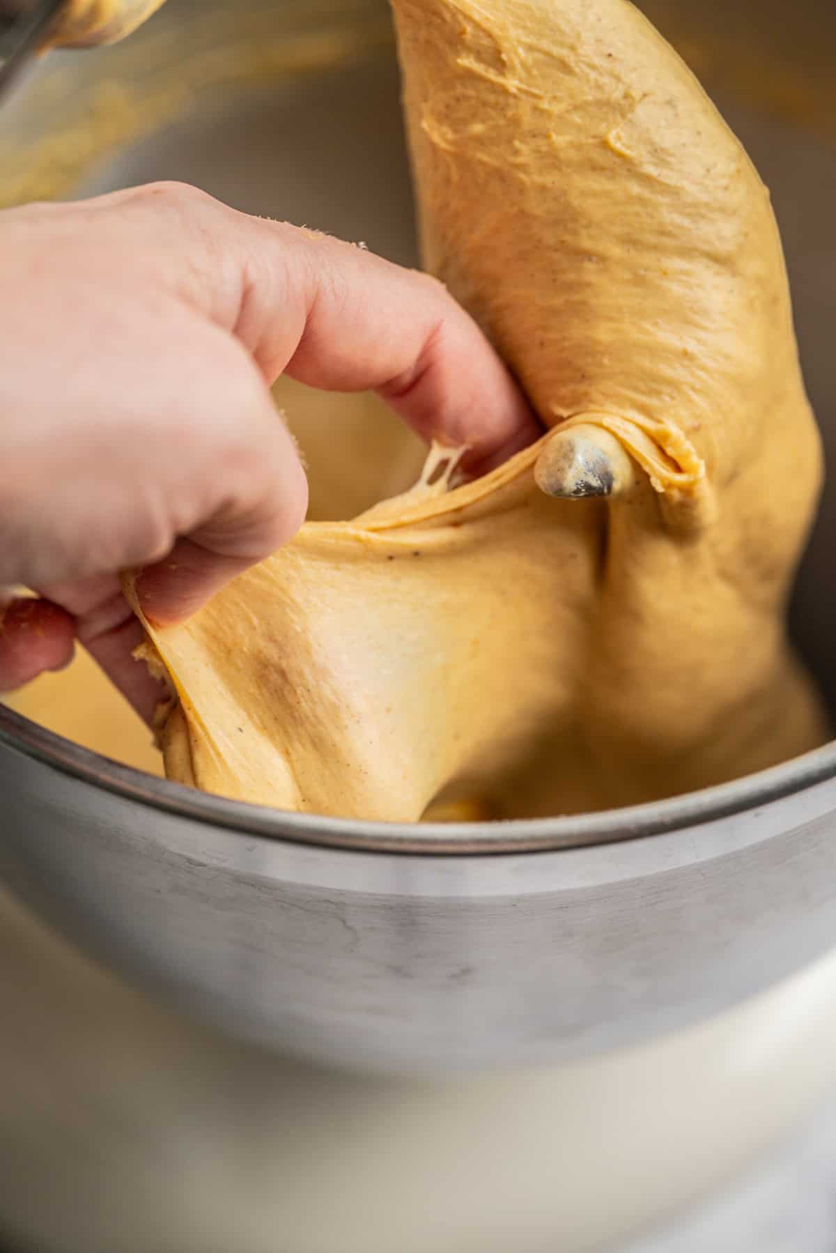 A hand stretches a smooth, elastic dough inside a metal mixing bowl, demonstrating its texture and gluten development.