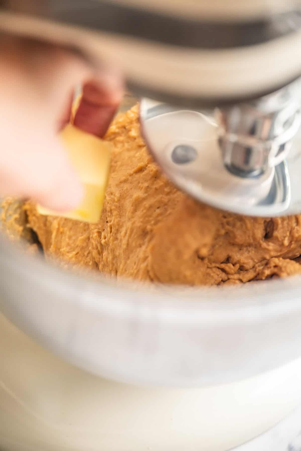 A close-up of a person adding a pat of butter to thick dough mixing in a stand mixer bowl.