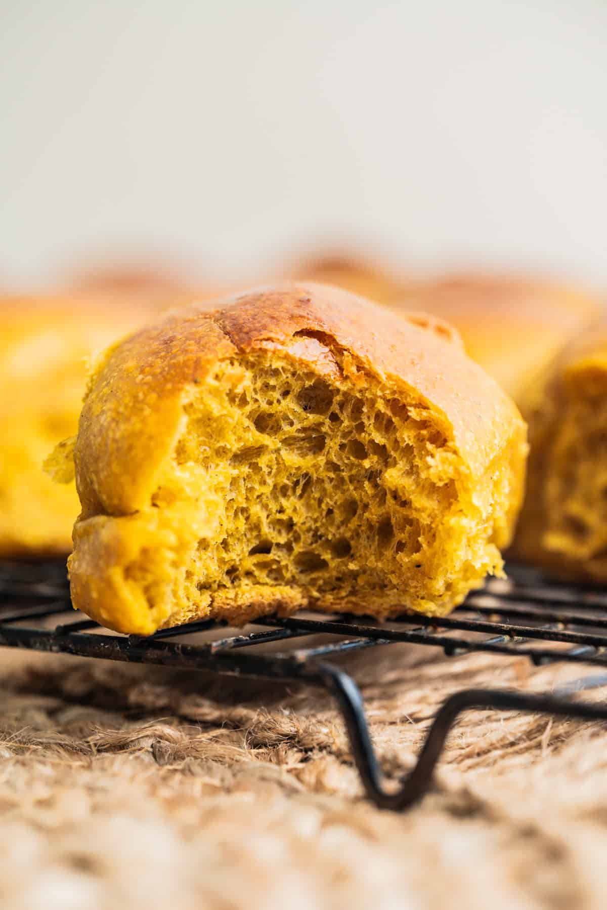 A close-up of a golden-brown pumpkin bread roll with a bite taken out, showing its soft and airy interior, resting on a black cooling rack.