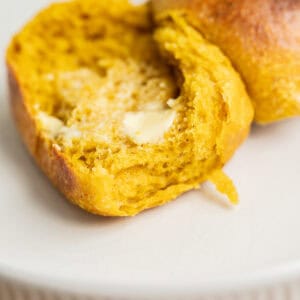 A close-up of a sourdough pumpkin dinner roll, split open on a white plate, with melted butter spread inside. The bread has a soft, fluffy texture and a slightly glossy crust.