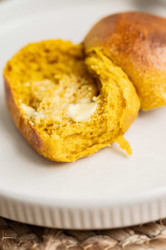 A close-up of a sourdough pumpkin dinner roll, split open on a white plate, with melted butter spread inside. The bread has a soft, fluffy texture and a slightly glossy crust.