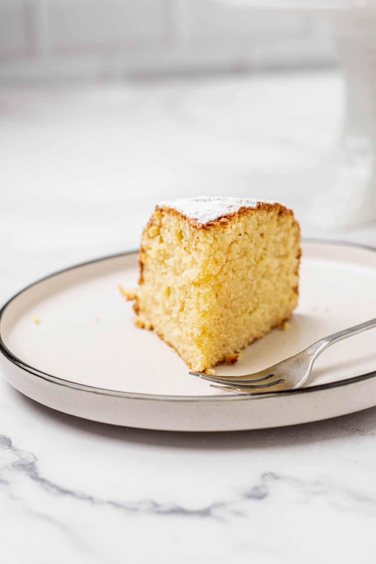 A single slice of yellow cake with a light dusting of powdered sugar on top sits on a round plate with a fork, on a marble surface.
