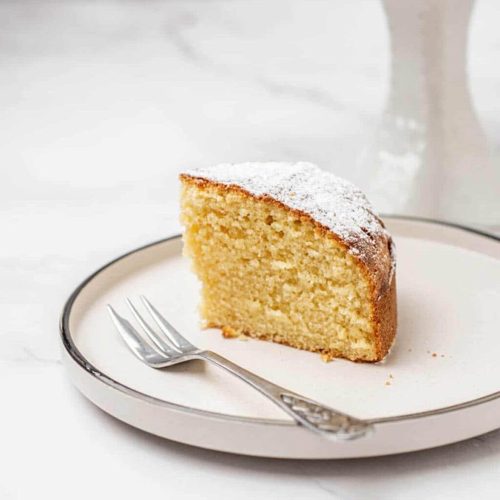 A single slice of yellow cake dusted with powdered sugar sits on a white plate with a fork beside it, against a light background.