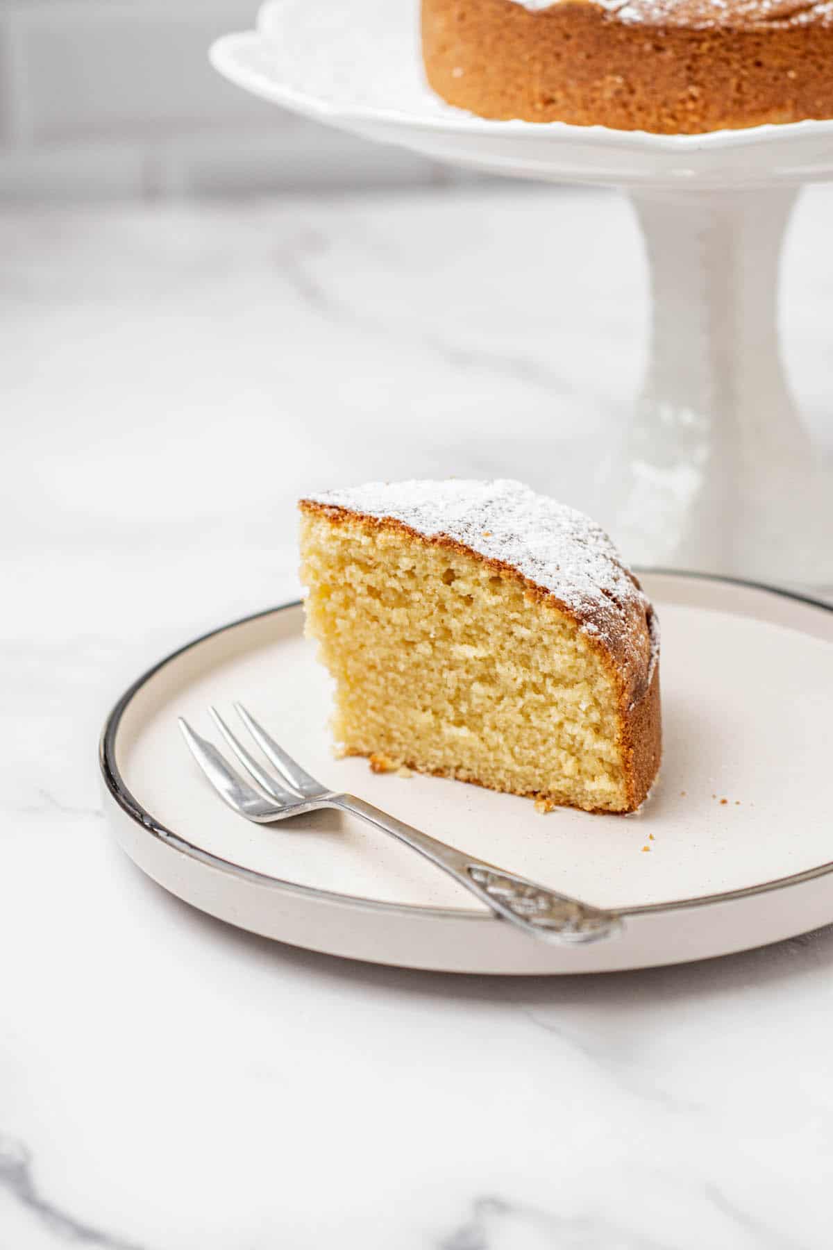 A single slice of yellow air fryer vanilla cake dusted with powdered sugar sits on a plate next to a fork, with the rest of the cake on a stand in the background.