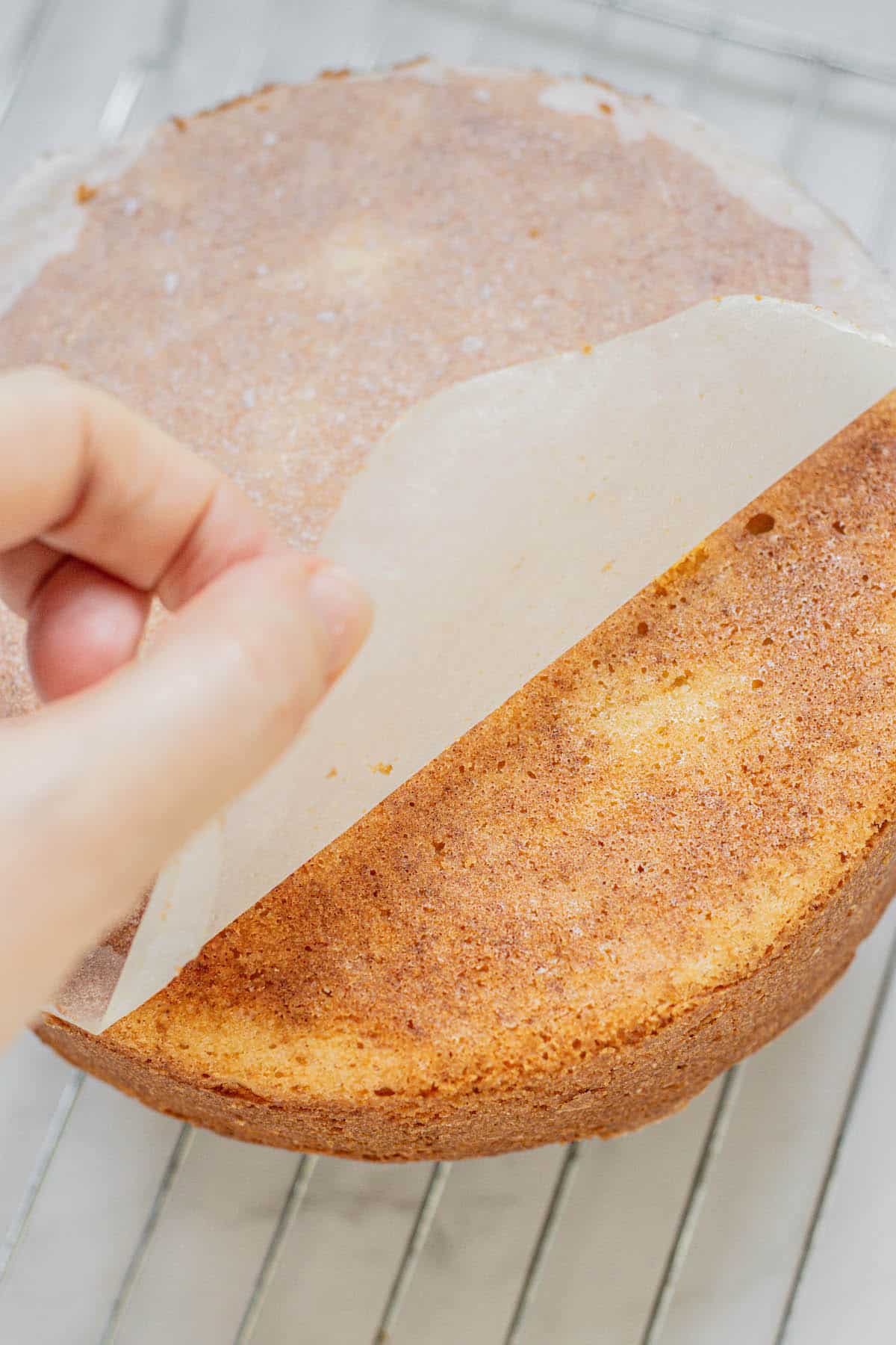 A hand lifts parchment paper off a freshly baked airfryer vanilla cake that is cooling on a wire rack.