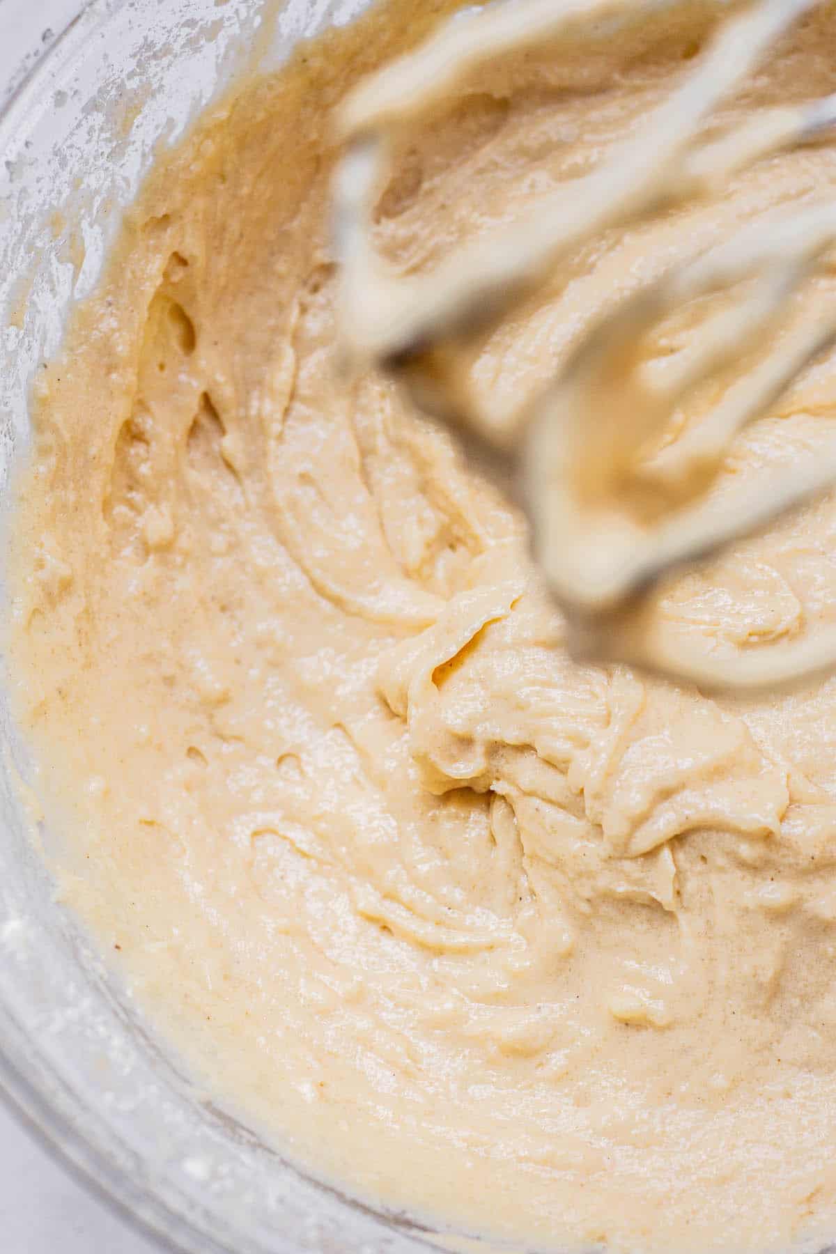 A close-up of creamy, light brown cake batter in a glass bowl with a hand mixer beater partially visible at the top right.