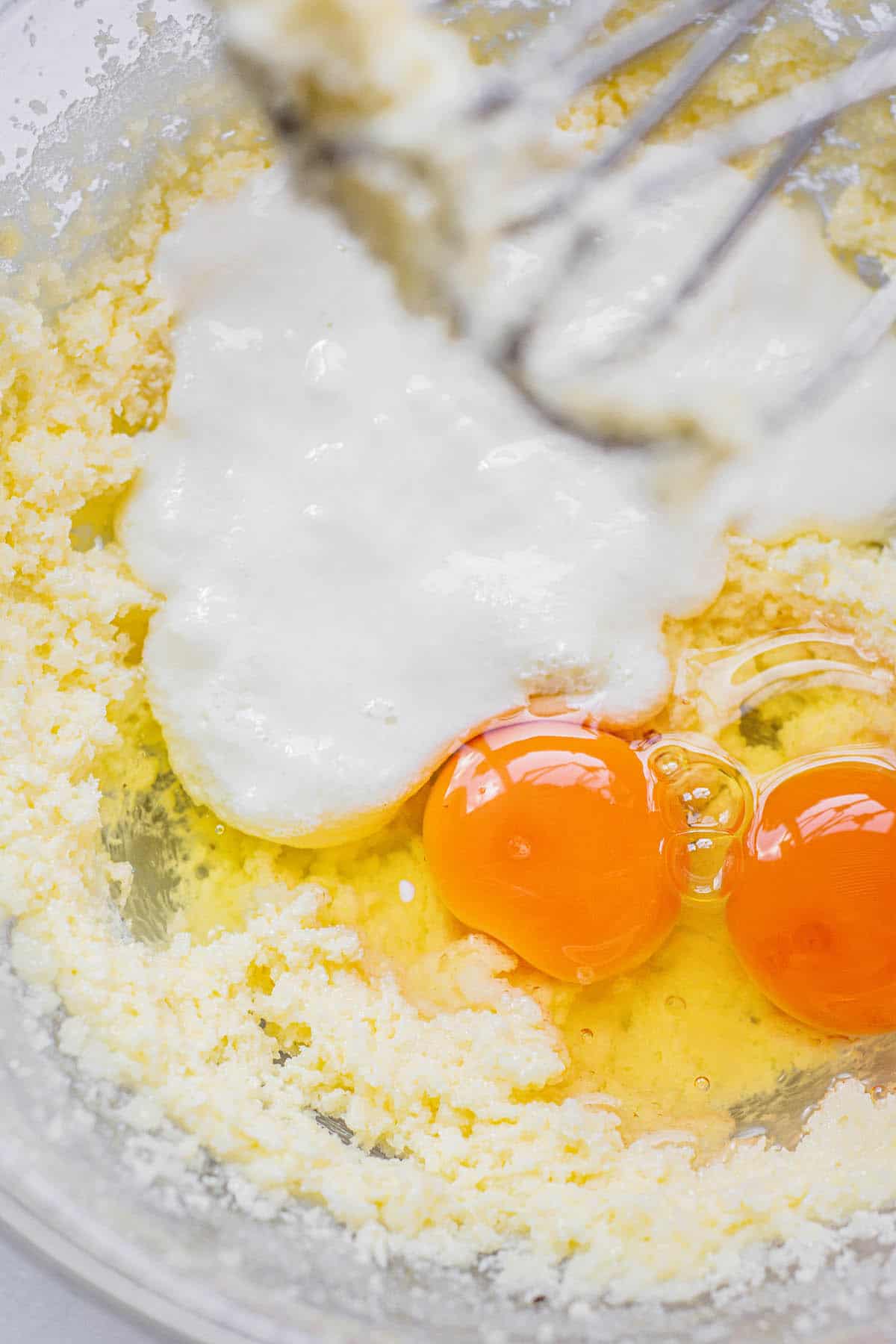 A close-up of a mixing bowl with two cracked eggs, creamed butter and sugar, and sour cream being mixed with a metal hand mixer.