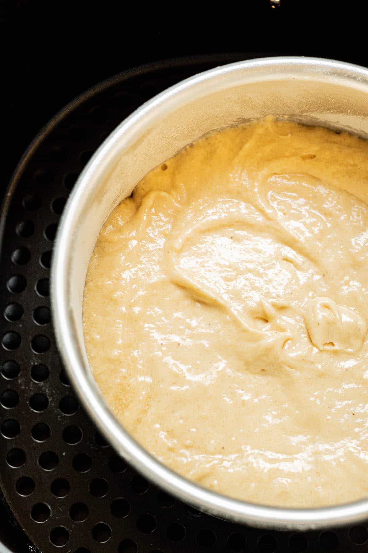 A round metal pan filled with smooth, pale yellow cake batter sits inside a black air fryer basket, ready to be baked.