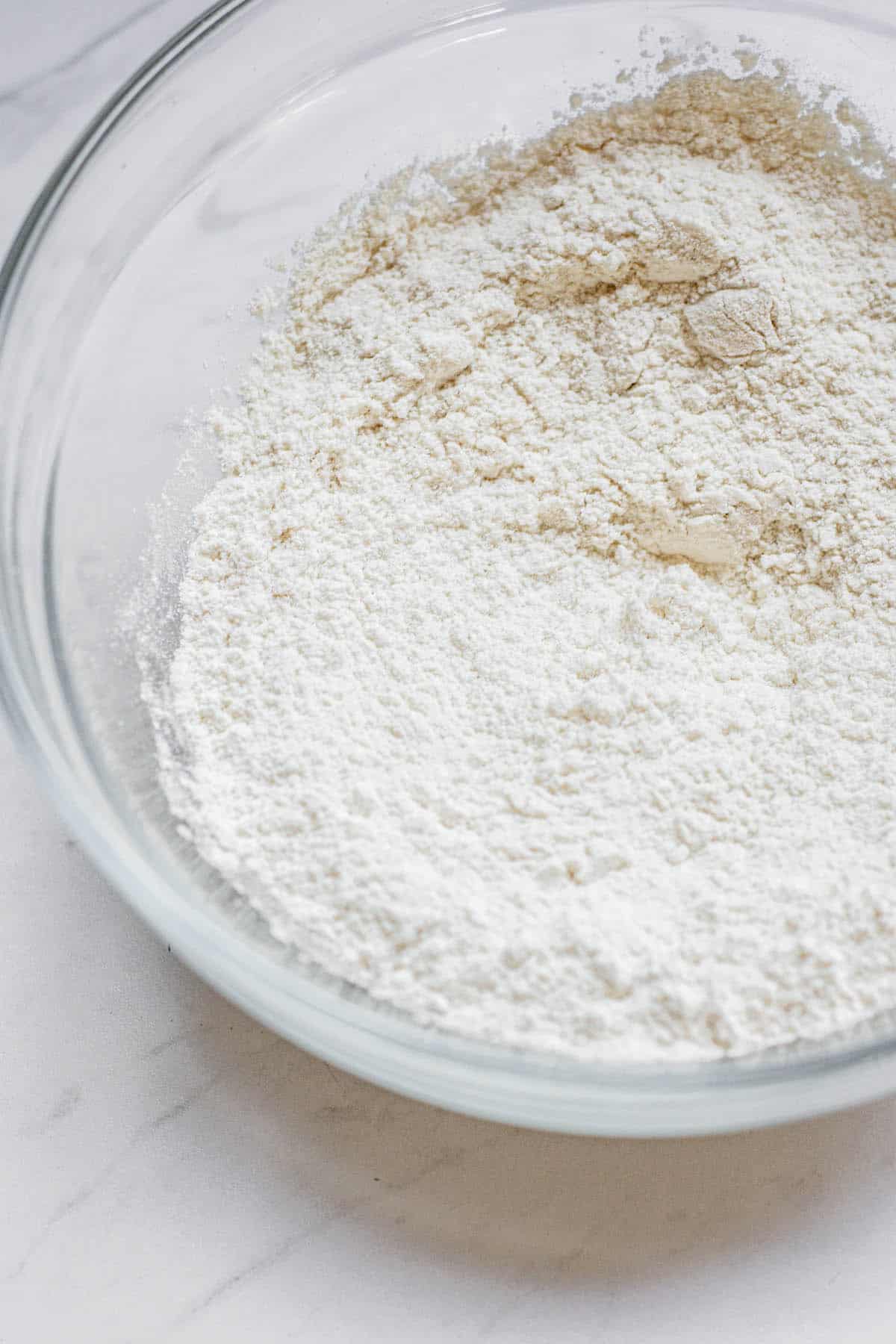 A clear glass bowl filled with white flour sits on a light-colored surface, showing a smooth and finely textured powder ready for baking or cooking.