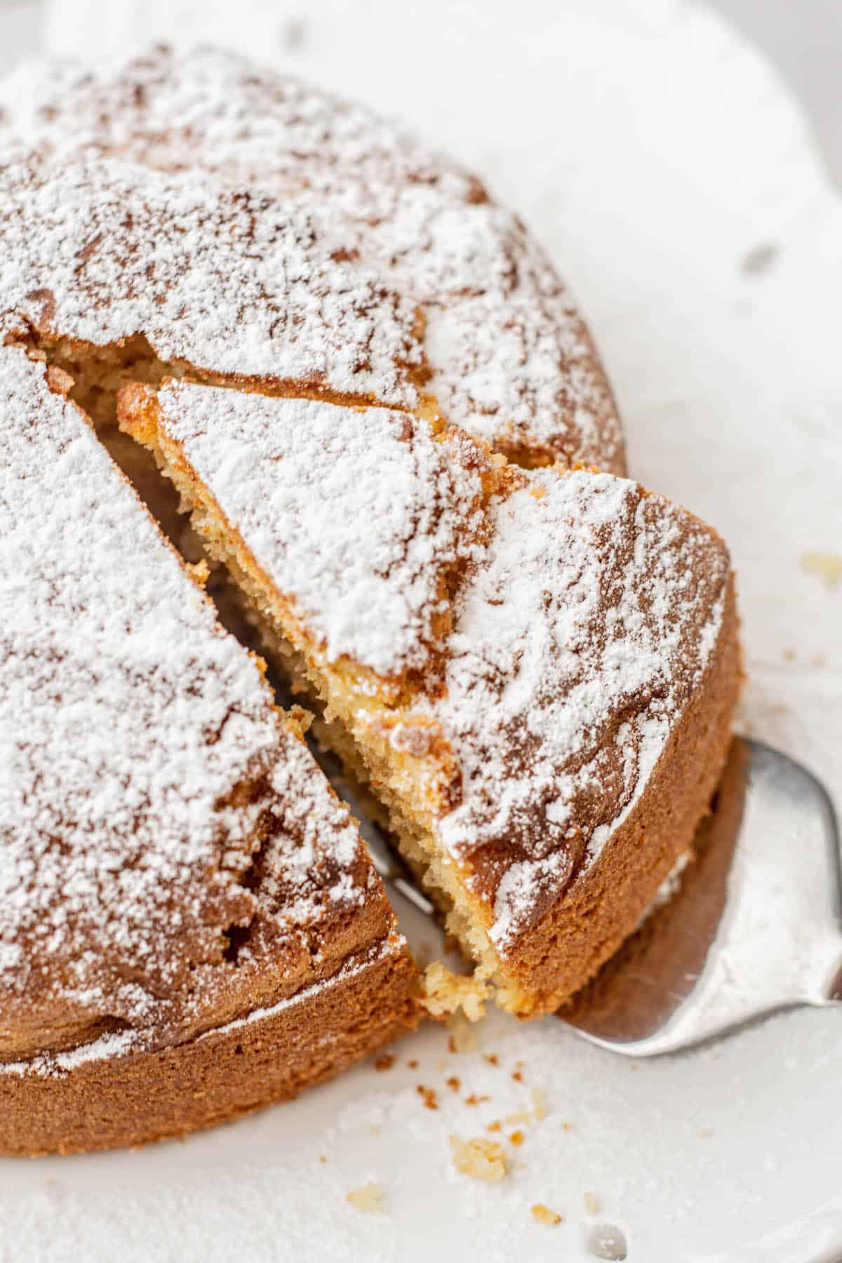 A round air fryer vanilla cake dusted with powdered sugar, with one slice being lifted out using a metal spatula. The cake is golden brown and sits on white parchment paper.