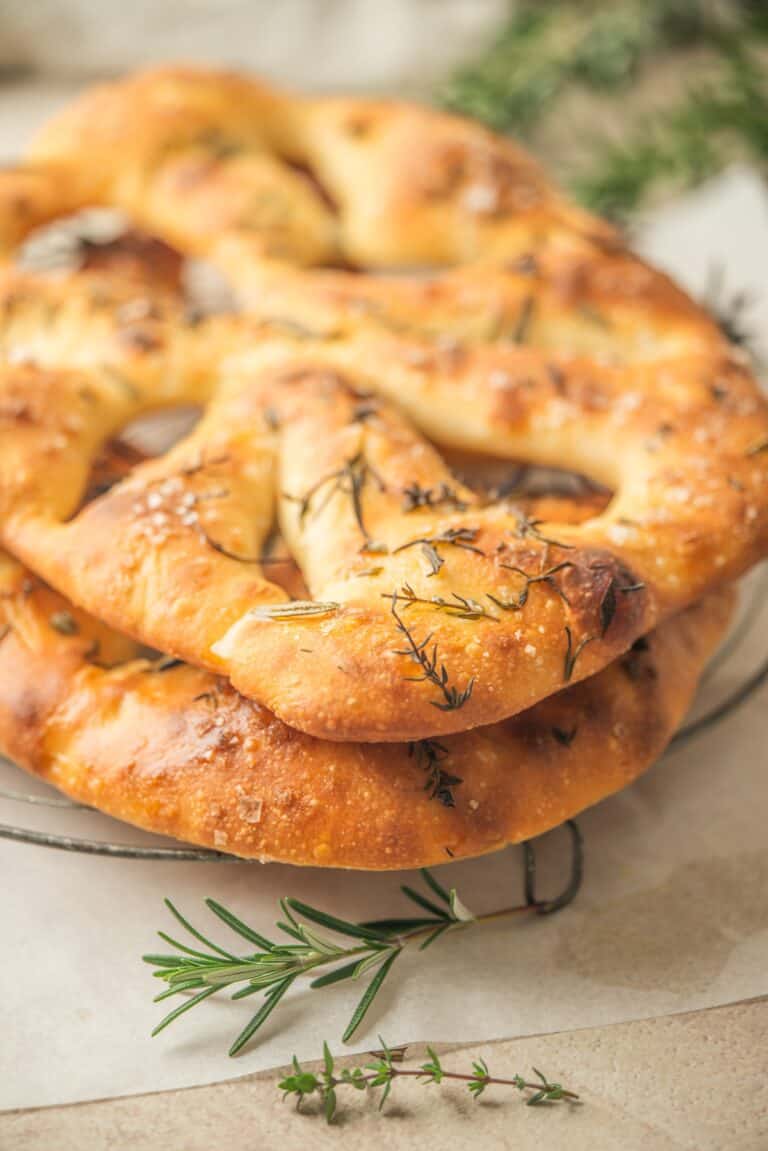 Three golden-brown sourdough fougasse flatbreads topped with fresh thyme and rosemary are stacked on a wire rack, with sprigs of rosemary in the foreground.